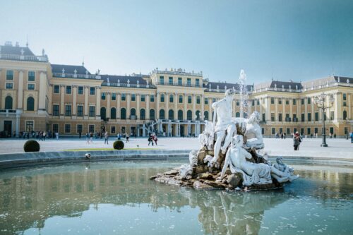 A fountain with statues in front of Schönbrunn Palace in Vienna, with people walking in the courtyard.