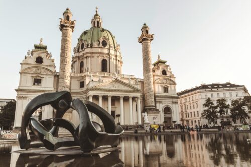 Charles Church in Vienna with its large green dome, two tall columns, and a sculpture reflected in the water in front.